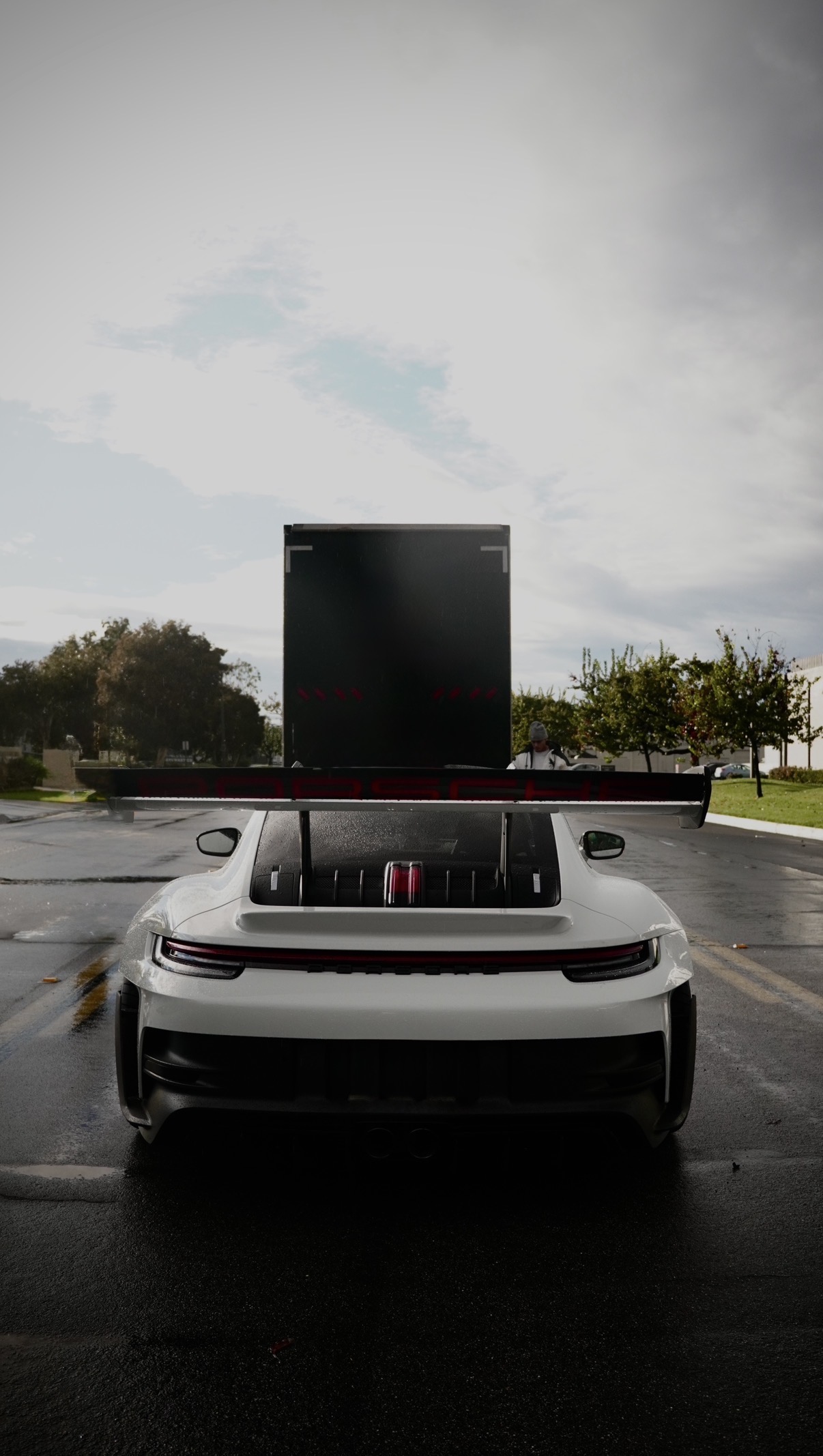 A white Porsche 911 GT3 RS seen head on from behind on a wet road, parked in front of an enclosed transport truck